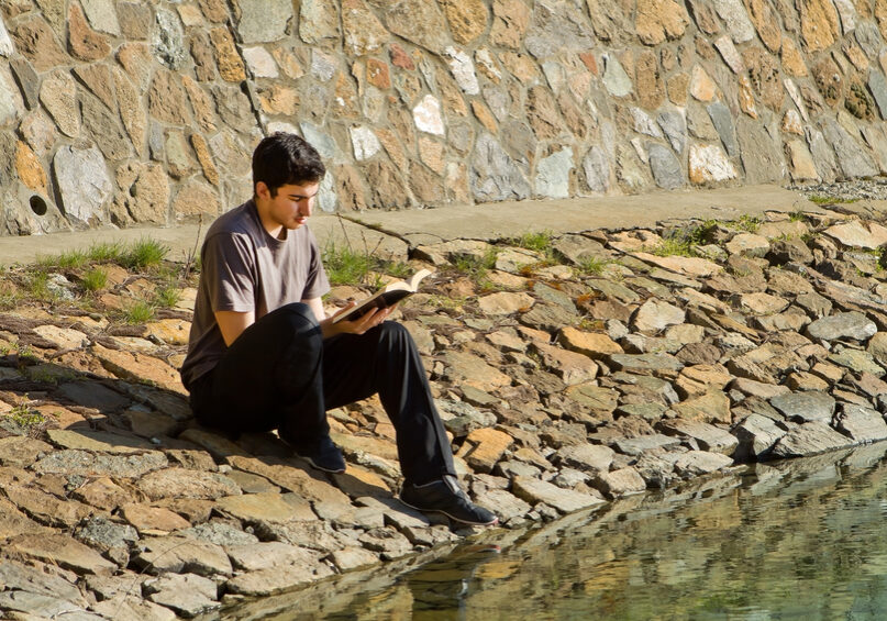 Young man sitting by a lake reading the Bible