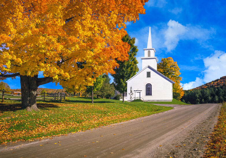 A large sugar maple in full autumn color and a country church fill the foreground leading back to the country side of the Green Mountains, Vermont